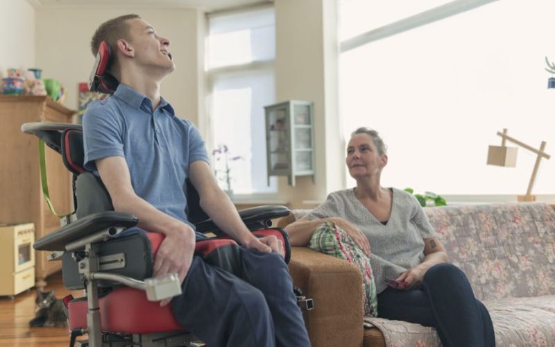 Color image of a real life young physically impaired ALS patient spending time with his mother at home. He is happy.